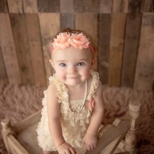 May include: A baby wearing a cream-colored lace dress and a pearl necklace. The baby is also wearing a pink headband with flower accents. The baby is in a small wooden bed.