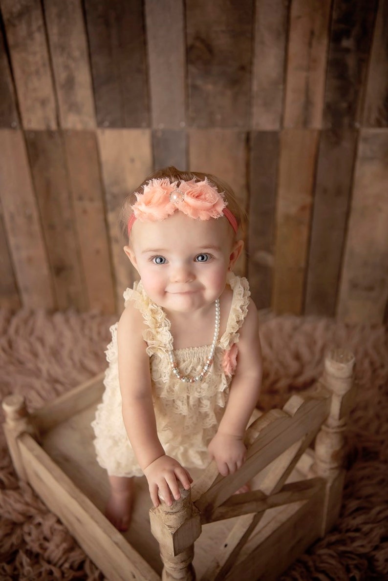 May include: A baby wearing a cream-colored lace dress and a pearl necklace. The baby is also wearing a pink headband with flower accents. The baby is in a small wooden bed.