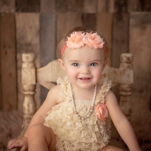 May include: A baby wearing a peach-colored floral headband and a cream-colored lace romper. A pearl necklace and a peach-colored flower accent the outfit. The baby is seated on a light-colored wooden chair against a rustic wooden backdrop.