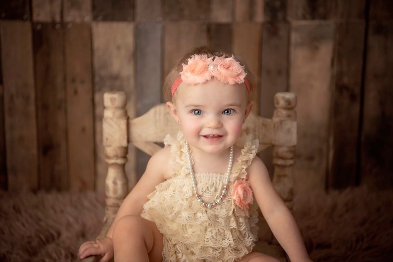 May include: A baby wearing a peach-colored floral headband and a cream-colored lace romper. A pearl necklace and a peach-colored flower accent the outfit. The baby is seated on a light-colored wooden chair against a rustic wooden backdrop.