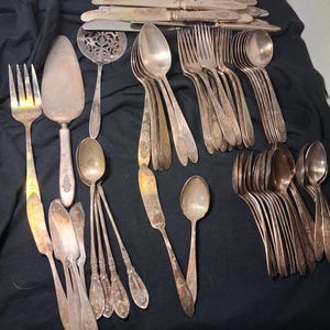 May include: A collection of vintage silverware, including forks, spoons, knives, and serving utensils. The silver-toned flatware is arranged on a dark surface, showcasing various designs and sizes, perfect for a formal dining setting.