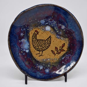May include: A handmade ceramic plate with a whimsical chicken design. The plate features a speckled blue and maroon rim, surrounding a tan center with a black chicken illustration and leaf sprig. A unique piece for kitchen decor.