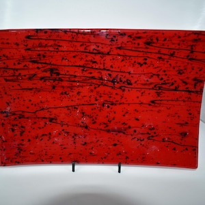 Puede incluir: Una bandeja decorativa rectangular de vidrio rojo con vetas y motas negras. La bandeja tiene una forma ligeramente curvada y se exhibe sobre un soporte de metal negro. El diseño es abstracto y moderno.