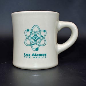May include: A cream-colored ceramic mug featuring a teal graphic of an atom symbol with a Zia sun symbol at its center. Below the graphic, the text reads Los Alamos NEW MEXICO in teal lettering. The mug has a sturdy handle and a smooth finish.