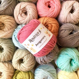 May include: A collection of yarn balls in various pastel and neutral colors. A central ball of dark pink yarn features a white paper label with the text Ros&aacute;rios 4 Amigomira Ecofriendly Collection and small illustrations of a bear, flowers, and hearts.