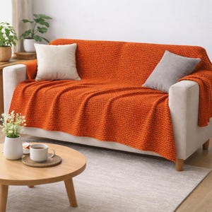 May include: An orange textured throw blanket covers a white sofa, with two throw pillows. A round wooden coffee table holds a cup of tea and a vase of flowers. The room has a neutral color palette.