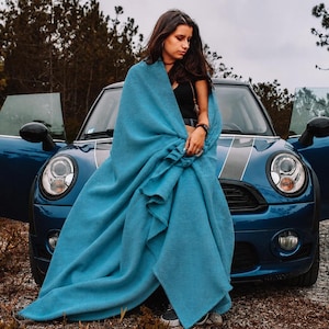 May include: A solid teal blue blanket draped over the front of a dark blue compact car with open doors. The blanket features a soft, textured fabric surface and is positioned to cover the front bonnet and grille area of the vehicle.