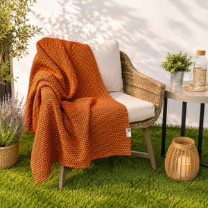 May include: An orange textured throw blanket draped over a wicker chair. The blanket has a dotted pattern and is on a green lawn. A small side table with a plant and a water bottle is next to the chair. The blanket is a warm, autumnal colour.