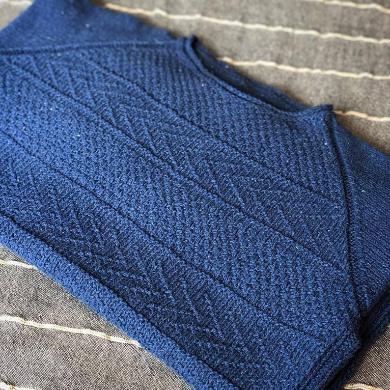 May include: A blue knitted sweater with a chevron pattern. The sweater is folded and laid on a striped fabric.