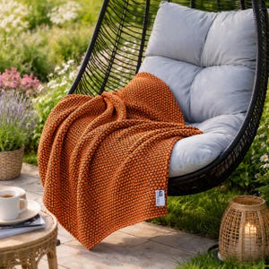 May include: An orange textured throw blanket draped over a gray cushion in a black wicker hanging chair. The blanket has a repeating dot pattern. A small table with a cup of coffee and a wicker lantern are in the background.