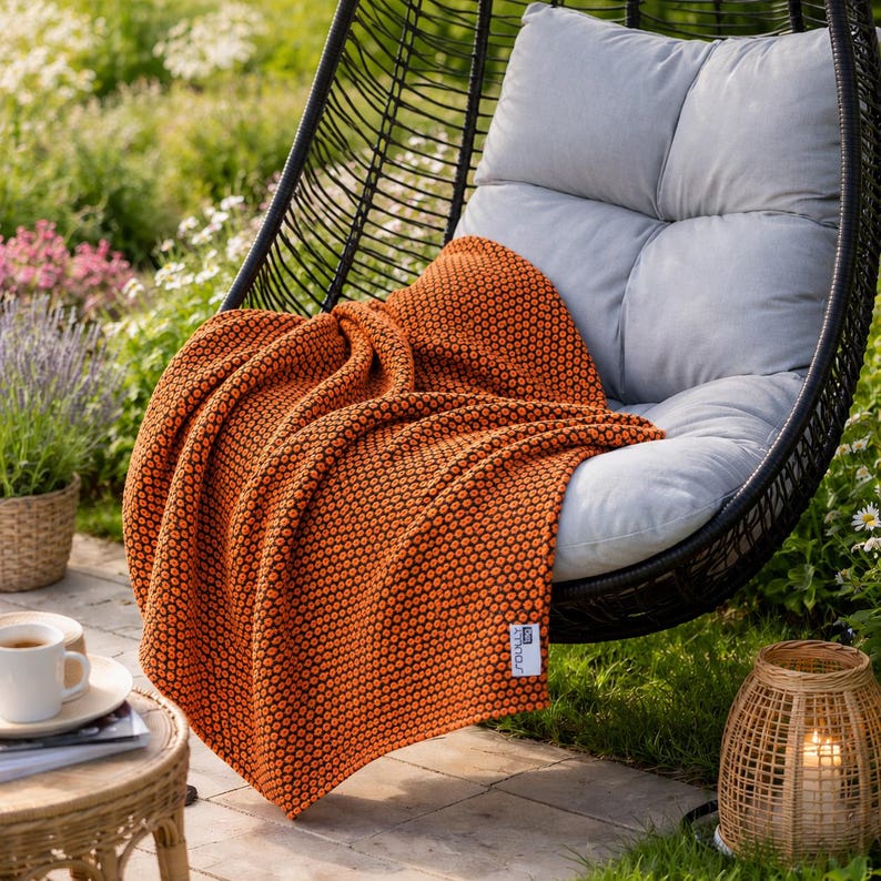 May include: An orange textured throw blanket draped over a gray cushion in a black wicker hanging chair. The blanket has a repeating dot pattern. A small table with a cup of coffee and a wicker lantern are in the background.