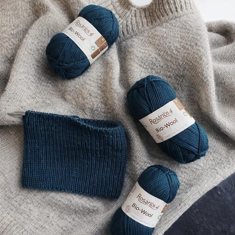 May include: Three skeins of dark teal Ros&aacute;rios 4 Bio-Wool yarn with white labels, accompanied by a rectangular dark teal knitted swatch on a light gray textured knit fabric background. The yarn labels feature the text Ros&aacute;rios 4 Bio-Wool.