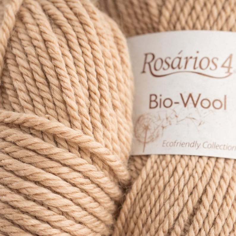 May include: A ball of light tan yarn with a white paper label featuring the text Ros&aacute;rios 4 Bio-Wool Ecofriendly Collection and a line drawing of a dandelion. The yarn has a thick, twisted texture and a soft, matte appearance.