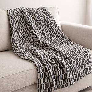 May include: A black and cream-colored throw blanket with a geometric pattern draped over a beige sofa. The blanket's design features a repeating pattern of vertical and horizontal lines, creating a textured appearance. The sofa has a neutral color and a simple design.
