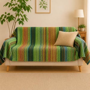 May include: A sofa covered with a striped throw blanket in shades of green, blue, and red. The blanket has vertical stripes. A beige cushion sits on the sofa. The sofa has wooden legs and is against a neutral background.
