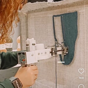 May include: A close-up of a white and silver machine embroidering a teal design on a beige fabric. The machine is held by a person's hand. The fabric is stretched on a frame. The image includes a heart icon with the number 169.