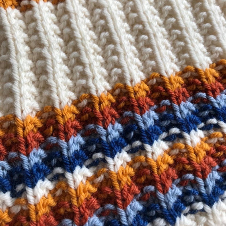 May include: A close-up of a knitted textile featuring a ribbed texture. The upper section is solid cream, while the lower section displays a repeating horizontal pattern of alternating stripes in mustard yellow, burnt orange, light blue, and navy blue.