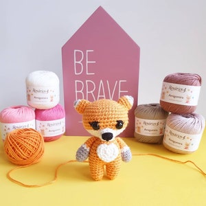 May include: A crocheted orange fox amigurumi with white accents, black eyes, and a black nose, positioned in front of a pink house-shaped sign with the text BE BRAVE, surrounded by several balls of Ros&aacute;rios 4 Amigomira yarn in various colors.