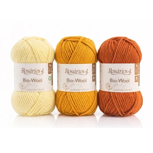 Pode incluir: Três novelos de fio Rosários 4 Bio-Wool em tons de amarelo, dourado e laranja enferrujado. Cada novelo de fio tem uma etiqueta branca com o nome da marca e detalhes do produto. O fio é bem enrolado, mostrando a textura da lã.