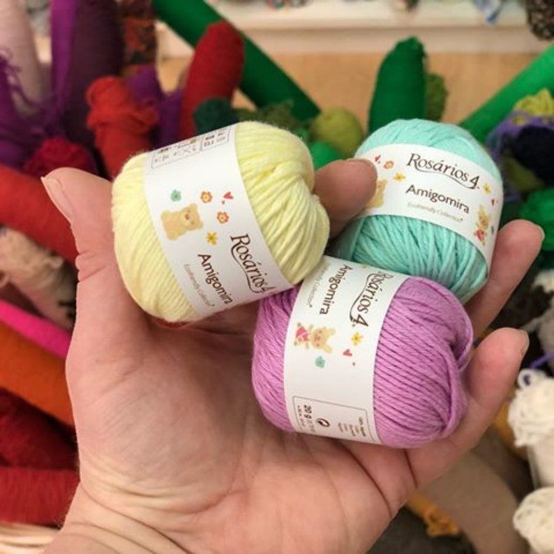 May include: Three small balls of Ros&aacute;rios 4 Amigomira yarn in pastel yellow, mint green, and light purple. Each ball features a white paper label with the brand name, product name, and small colorful illustrations of animals and flowers.