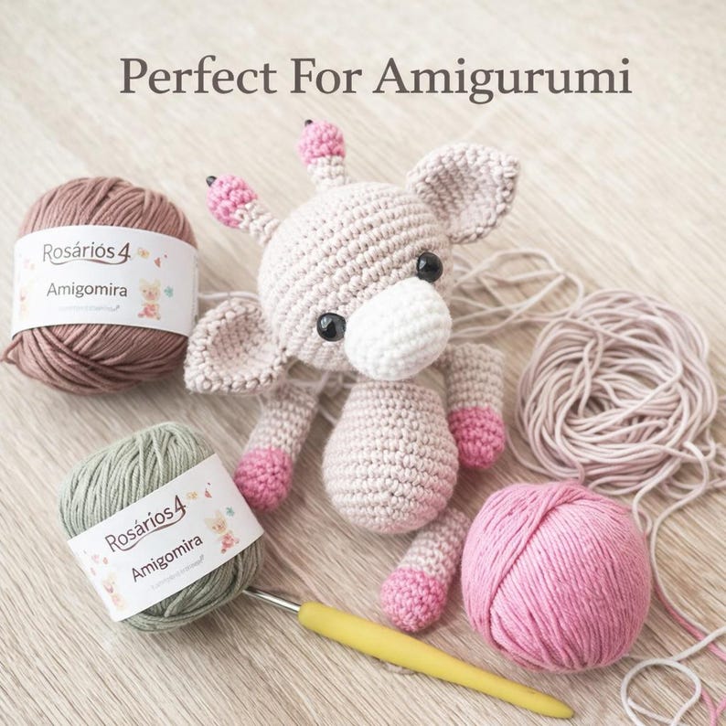 May include: A crocheted giraffe toy with pink accents, two balls of Ros&aacute;rios 4 Amigomira yarn in dusty rose and sage green, a loose ball of light pink yarn, and a yellow crochet hook. The text Perfect For Amigurumi is visible at the top.