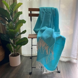 Może przedstawiać: Turkusowy koc z białymi frędzlami, drapowany na drewnianym krześle. Koc jest miękki i przytulny, idealny do dodania odrobiny koloru do każdego pomieszczenia.