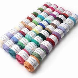 May include: A collection of small yarn balls arranged in a grid, featuring a wide variety of solid colors including red, blue, green, yellow, purple, and orange. Each ball is wrapped with a white paper label printed with the text Ros&aacute;rios 4 Amigomira.