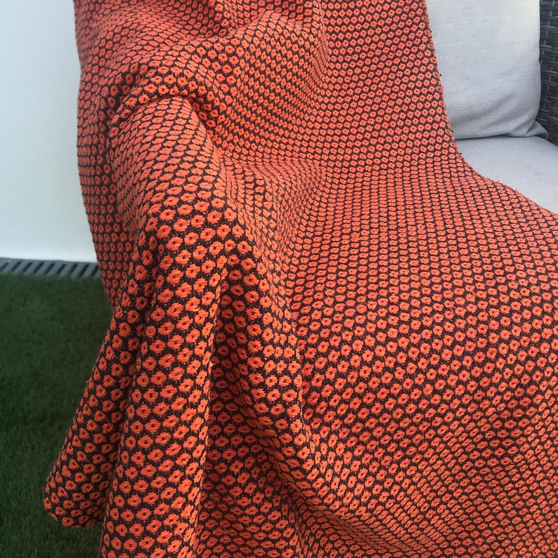 May include: An orange and black patterned throw blanket draped over a chair. The blanket features a repeating floral design. The color palette is a warm orange and black, creating a cozy and inviting aesthetic. The blanket is made of a soft, textured material.