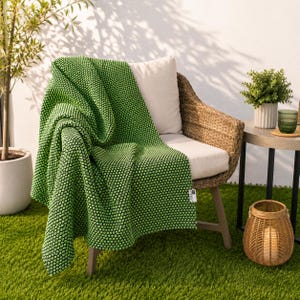 May include: A green textured throw blanket draped over a wicker chair with a white cushion. The blanket has a dotted pattern. A small side table with a plant and glass is next to the chair. A decorative basket sits on the ground.