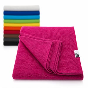 May include: A folded magenta blanket with a textured weave, displaying a tag with text. A stack of folded blankets in various colors, including white, blue, green, and yellow, is visible in the background. The blankets appear to be made of a soft material.