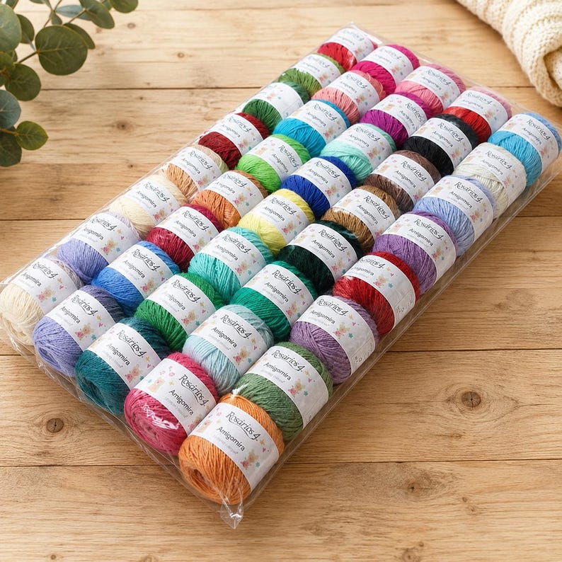 May include: A collection of small, round yarn balls in a wide variety of colors, including red, blue, green, yellow, purple, and orange. Each ball is wrapped in a white paper label with the text Rosarios 4 Amigomira, arranged in a clear plastic tray.