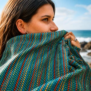 May include: A colorful woven textile with a repeating pattern of blue, orange, yellow, and green squares. The textile is draped over a person's face, with the ocean visible in the background. The textile is likely a blanket or shawl.