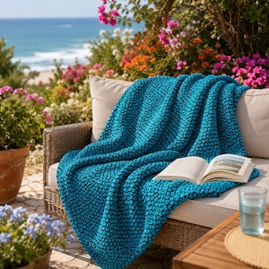 May include: A teal blue textured throw blanket on a wicker chair with a beige cushion. An open book and a glass of water are on a wooden table. The background shows the sea and colourful flowers.