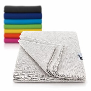 May include: A folded light gray towel with a textured surface, with a stack of colorful towels in the background. The folded towel has a small white tag with black text. The stack of towels includes black, blue, green, yellow, orange, and pink.