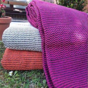 Puede incluir: Tres mantas plegadas en fucsia, blanco y negro, y naranja. Cada manta presenta un patrón de chevrón. Las mantas están apiladas sobre hierba, lo que sugiere su uso al aire libre. Probablemente hechas de material suave.