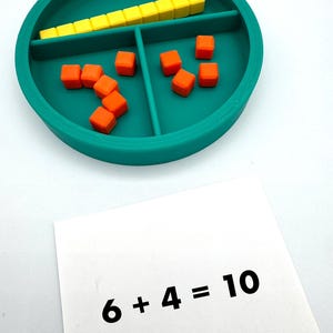 May include: A teal circular divided tray containing a row of ten yellow cubes and several orange cubes, accompanied by a white card featuring the printed black text 6 + 4 = 10. The tray is segmented into two main sections for sorting small objects.