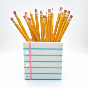 May include: A white pencil holder shaped like a piece of notebook paper with horizontal blue lines and a vertical pink margin line, filled with a collection of yellow wooden pencils featuring metallic gold ferrules and pink erasers at the top.
