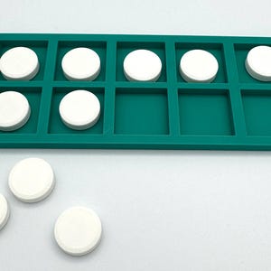 May include: A teal rectangular ten frame tray with the text Ten Frame printed on the left side. The tray contains seven white circular tokens placed in the grid squares, with three additional white circular tokens resting outside the tray.