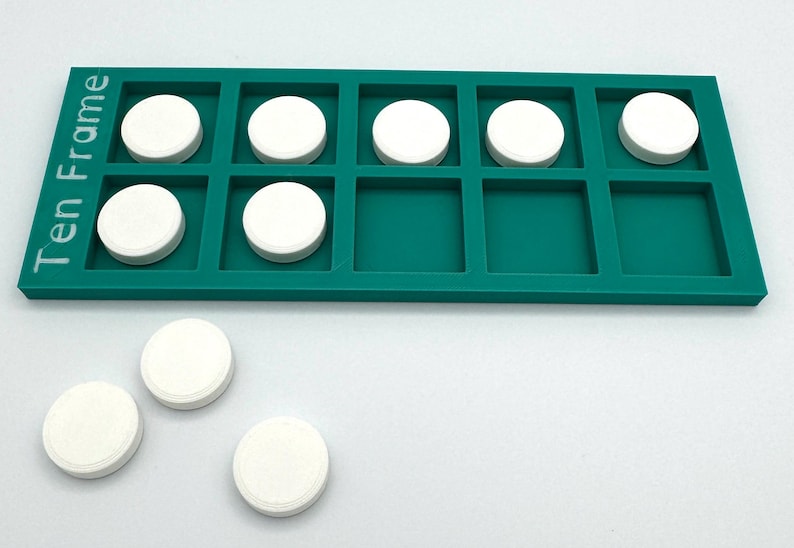 May include: A teal rectangular ten frame tray with the text Ten Frame printed on the left side. The tray contains seven white circular tokens placed in the grid squares, with three additional white circular tokens resting outside the tray.
