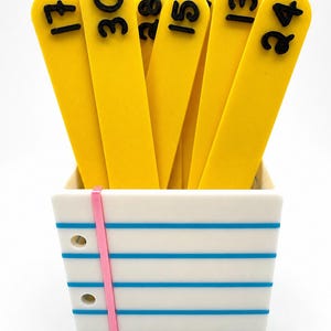 May include: A white square container designed to look like a piece of notebook paper with blue horizontal lines and a pink vertical margin line, holding several bright yellow flat sticks with raised black numbers 17, 30, 28, 15, 13, and 24.