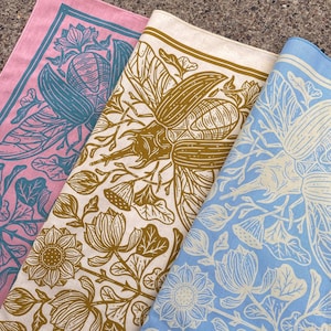 May include: Three fabric squares with different patterns. The top square is pink with a teal floral design. The middle square is cream with a brown floral design featuring a large beetle. The bottom square is light blue with a white floral design.