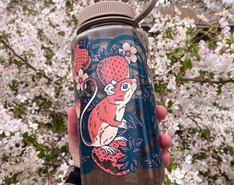May include: A clear water bottle with a tan lid and handle, featuring a whimsical design of a pink mouse, strawberries, and flowers. The bottle is filled with a brown liquid and is held against a backdrop of white blossoms.