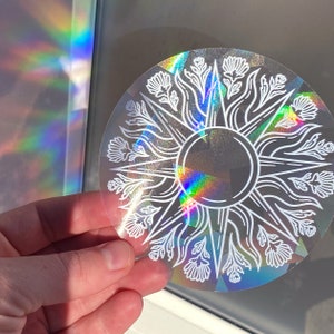 May include: A clear, circular sticker with a white sunburst design featuring floral elements. The sticker has a holographic finish, reflecting a rainbow of colors. The design includes a central circle and radiating lines.