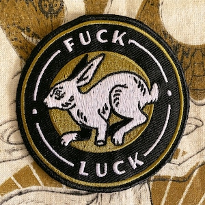 May include: A black and gold round embroidered patch with a white rabbit running inside a circle. The text "FUCK LUCK" is written in white around the circle.