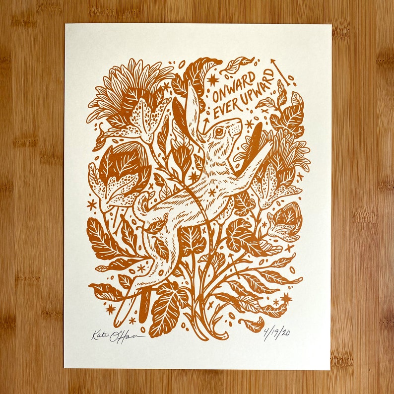 Pu&ograve; includere: Una stampa linoleografica marrone e bianca di un coniglio che salta in alto attraverso un campo di fiori. La stampa &egrave; intitolata "Onward Ever Upward" ed &egrave; firmata dall'artista, Katie O'Hara.