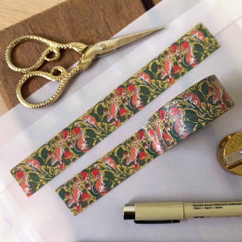 May include: Two rolls of washi tape with a repeating pattern of red and green mice on a yellow background. The tape is on a white surface with a pair of gold scissors and a pen.