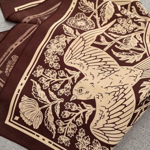 May include: A brown fabric bandana with a beige bird and floral design. The bird is flying with its wings spread and has a detailed beak and eye. The floral design includes leaves and flowers.