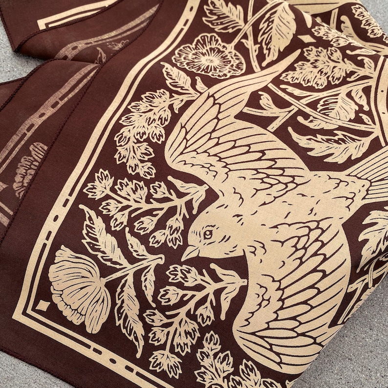May include: A brown fabric bandana with a beige bird and floral design. The bird is flying with its wings spread and has a detailed beak and eye. The floral design includes leaves and flowers.