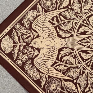 May include: A brown bandana with a white bird and floral design. The bird is flying with its wings spread out. The flowers are in bloom and have intricate details.