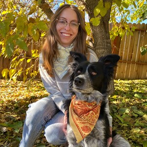 May include: A woman with glasses and a blue bandana is holding a black and white dog with a brown and orange bandana. The dog is wearing a bandana with a floral pattern.